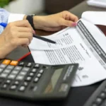 Seseorang menganalisis dokumen keuangan dengan kalkulator dan pena di atas meja